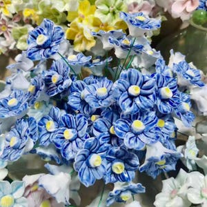 May include: A close-up of a bouquet of ceramic flowers. The flowers are mainly blue with white and yellow centres. Other flowers in the background are yellow, white, and pink. The flowers have green stems.