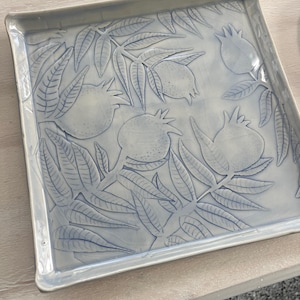 May include: A square ceramic serving tray with a blue glaze and a raised design of leaves and pomegranates.
