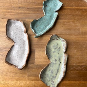 May include: Three ceramic dishes shaped like the state of New Jersey. The dishes are glazed in different colors: light blue, white, and light green. Each dish has a speckled finish.