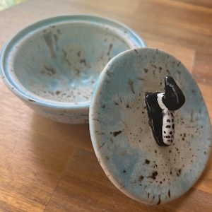 Könnte beinhalten: Keramikschale mit Deckel, mit hellblauer und braun gesprenkelter Glasur. Der Deckel hat einen schwarz-weißen dekorativen Griff. Die Schale ist offen und zeigt ihr Inneres. Schale und Deckel stehen auf einer Holzoberfläche.