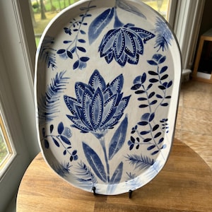 May include: A white oval ceramic serving tray with a blue floral design. The tray has a blue floral pattern with white accents and is displayed on a wooden stand.
