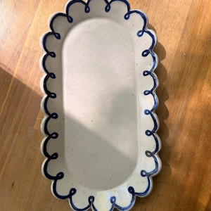 May include: A speckled, off-white ceramic serving platter with a scalloped edge and a dark blue wavy border. The oval-shaped dish is set on a wooden surface. This decorative platter is ideal for serving appetizers or desserts.