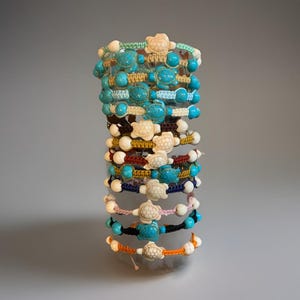 May include: A stack of handmade bracelets featuring turtle charms and turquoise beads. The bracelets are made with various colored cords, including white, blue, brown, and orange. Each bracelet has a unique design and color combination.