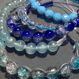 May include: A collection of beaded bracelets in shades of blue and clear. The bracelets feature various bead shapes, including faceted and round, with a mix of solid and crackled glass beads. The bracelets are strung on cords, showcasing a range of light to dark blue hues.