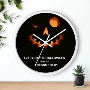 Op de afbeelding: Ronde witte wandklok met een zwarte wijzerplaat. De wijzerplaat heeft een oranje gesneden pompoenontwerp en witte wijzers. De tekst luidt: "EVERY DAY IS HALLOWEEN isn't it? FOR SOME OF US".