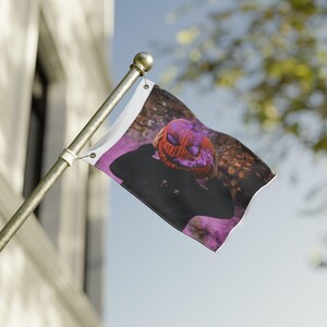 Op de afbeelding: Kleine vlag met een persoon met een gesneden pompoenhoofd. De pompoen is oranje en paars, tegen een zwarte achtergrond. De vlag is bevestigd aan een gouden paal en wappert in de wind.