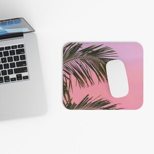 Op de afbeelding: Een rechthoekige muismat met een roze en paarse gradiënt achtergrond. De muismat heeft een groen palmbladontwerp en een witte computermuis. Een zilveren laptop staat in de linkerbovenhoek.
