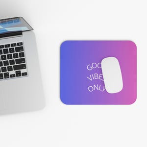 Op de afbeelding: Een rechthoekige muismat met een paarse en blauwe gradiënt, met de witte tekst "GOOD VIBE ONLY". Een witte computermuis ligt op de mat, naast een laptop.
