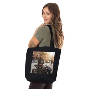 May include: Black tote bag with a photograph of a person on a black motorcycle. The bag has black straps and is held by a person wearing a green shirt.