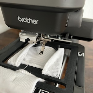 Könnte beinhalten: Eine Brother-Stickmaschine mit einem weißen Stoffring. Die Maschine ist gerade dabei, ein Design auf den Stoff zu sticken. Die Maschine hat eine schwarz-graue Farbgebung und das Wort "brother" ist auf der Vorderseite der Maschine aufgedruckt.