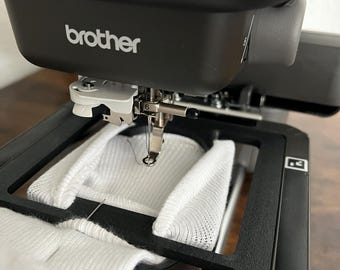 Può includere: Una macchina da ricamo Brother con un telaio in tessuto bianco in posizione. La macchina sta ricamando un disegno sul tessuto. La macchina ha una combinazione di colori nero e grigio e la parola "brother" è stampata sulla parte anteriore della macchina.