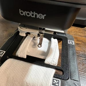 Könnte beinhalten: Eine schwarze Brother-Stickmaschine mit einer weißen Socke, die im Rahmen befestigt ist. Die Nadel der Maschine befindet sich über dem Stoff und ist bereit zum Sticken. Die Maschine steht auf einer Holzoberfläche.