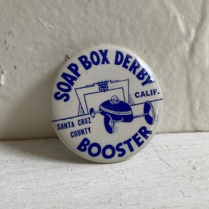 May include: A vintage white button with blue text and graphics. The button reads "SOAP BOX DERBY CALIF." and "SANTA CRUZ COUNTY BOOSTER." A blue race car graphic is in the center. The button is round and appears to be made of metal.