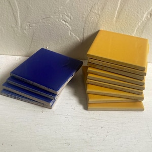 May include: Stacks of square ceramic tiles in two colors: deep blue and golden yellow. The tiles are arranged in small stacks, showcasing their smooth, glossy surfaces. The image highlights the contrast between the two colors.