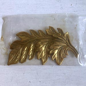 May include: A gold-colored metal leaf-shaped decorative element, possibly a furniture applique or embellishment, is sealed in a clear plastic bag. The leaf has detailed veining and a slightly aged appearance, suggesting a vintage or antique style.