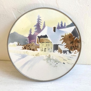 Puede incluir: Plato decorativo redondo con una escena invernal. El plato presenta una casa cubierta de nieve, árboles y un camino, todo sobre un cielo amarillo y púrpura. El plato tiene un borde de metal plateado.