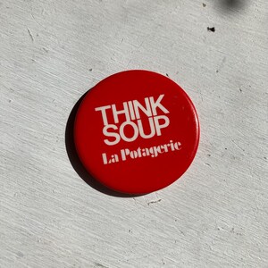 May include: A round, red button with the words "THINK SOUP" in white block letters above "La Potagerie". The button is a simple, eye-catching design.