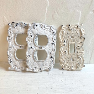 May include: Three off-white light switch plates with a distressed finish and ornate, raised scrollwork borders. Two plates feature double circular openings, while the third plate has a single rectangular opening, all with a textured grid pattern.