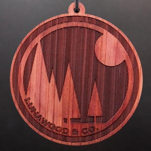 May include: A round, wooden ornament with a red-brown color. It features a stylized design of three trees and a crescent moon, with the text "LUNAWOOD & CO." inscribed around the edge.