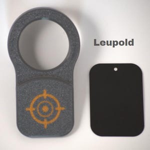 May include: A gray, textured accessory with a circular handle and a rectangular base featuring an orange target design. A separate black, rectangular piece with a small hole is also present. The word "Leupold" is printed on the image.