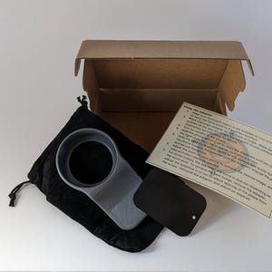 May include: A gray phone ring holder with a black adhesive disc and instructions for using the adapter. The holder is in a black velvet pouch and a brown cardboard box.