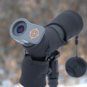 May include: A black spotting scope with a gray eyepiece featuring an orange target logo. The scope is mounted on a black tripod and has a lens cap attached. The background is blurred, suggesting an outdoor setting.