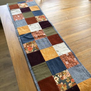 May include: A rectangular table runner constructed from a patchwork of square fabric pieces in various colors and textures, including solid rust, olive green, navy blue, mustard yellow, and dark purple, alongside floral and botanical patterns.