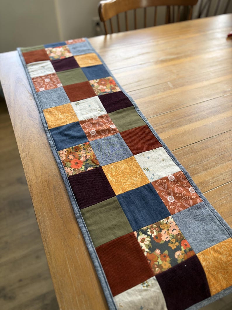 K&ouml;nnte beinhalten: Ein rechteckiger Tischl&auml;ufer aus einem Patchwork aus quadratischen Stoffst&uuml;cken in verschiedenen Farben und Texturen, darunter einfarbiges Rostrot, Olivgr&uuml;n, Marineblau, Senfgelb und Dunkellila sowie florale und botanische Muster.