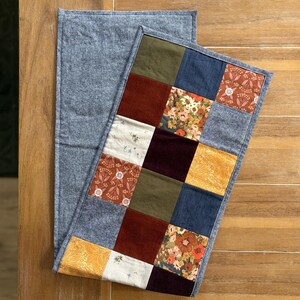 K&ouml;nnte beinhalten: Ein rechteckiger Tischl&auml;ufer mit einem Patchwork-Design aus verschiedenen Stoffquadraten in Olivgr&uuml;n, Senfgelb, Dunkelrot, Marineblau und Creme, akzentuiert durch florale Muster und einfarbige Texturen, eingefasst mit einem grauen Rand.