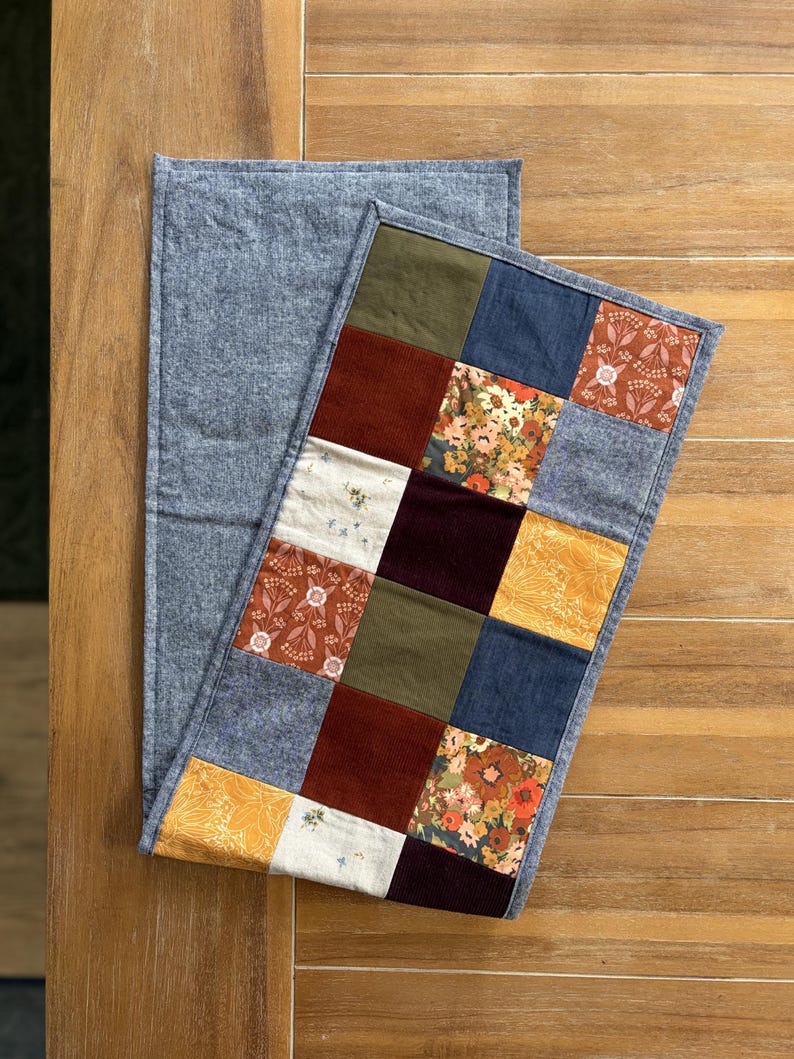 K&ouml;nnte beinhalten: Ein rechteckiger Tischl&auml;ufer mit einem Patchwork-Design aus verschiedenen Stoffquadraten in Olivgr&uuml;n, Senfgelb, Dunkelrot, Marineblau und Creme, akzentuiert durch florale Muster und einfarbige Texturen, eingefasst mit einem grauen Rand.