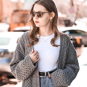 Puede incluir: Una mujer con un cárdigan gris de punto trenzado sobre una camiseta blanca y vaqueros azules. También lleva gafas de sol y un cinturón negro. El cárdigan tiene cierre de botones y un diseño texturizado.