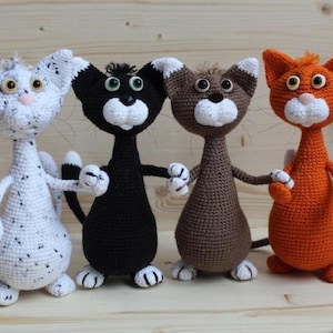 Może przedstawiać: Cztery ręcznie robione szydełkowe lalki kotów w kolorze białym, czarnym, brązowym i pomarańczowym. Każdy kot ma biały pyszczek, łapy i uszy. Koty stoją obok siebie, trzymają się za ręce i mają haftowane rysy twarzy.