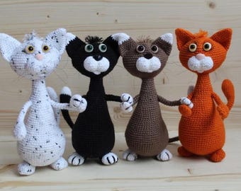 May include: Four handmade crochet cat dolls in white, black, brown, and orange. Each cat has a white muzzle, paws, and ears. The cats are standing side-by-side, holding hands, and have embroidered facial features.