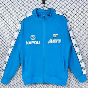 Könnte beinhalten: Hellblaue Jacke mit Reißverschluss und weißen Details an den Ärmeln. Die Jacke zeigt das Wort "NAPOLI" und das Wort "Mars" in Weiß. Die Ärmel haben ein sich wiederholendes weißes Logomuster auf einem weißen Streifen.