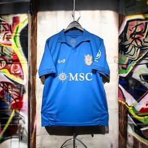 May include: A bright blue short-sleeved sports jersey with a polo collar. The front features the white text MSC with a star logo, the EA7 logo, and a crest with the letter N. The jersey is displayed on a black hanger against a light wall.