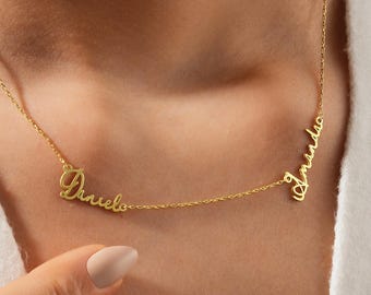 May include: A gold-colored necklace featuring two names in a cursive font. The names "Danielo" and "Amanda" are connected by a delicate chain. The necklace is displayed on a person wearing a white top.