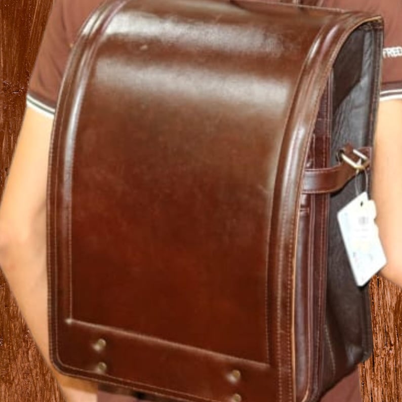 Może przedstawiać: Brązowa sk&oacute;rzana torba szkolna o prostokątnym kształcie i zaokrąglonej g&oacute;rze. Torba ma zapięcie na klapę, uchwyt i klamrę. Torba jest trzymana przez osobę.