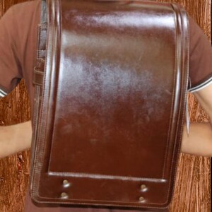 Może przedstawiać: Brązowy sk&oacute;rzany plecak szkolny o prostokątnym kształcie. Torba posiada klapę, pasek na ramię i dwa małe metalowe nity na dole. Sk&oacute;ra ma błyszczące wykończenie.