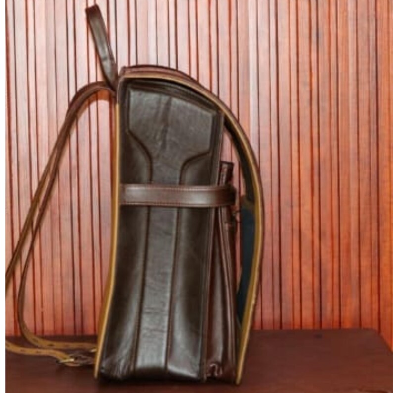 Może przedstawiać: Ciemnobrązowy sk&oacute;rzany plecak o strukturalnym designie. Torba posiada g&oacute;rny uchwyt, regulowane paski na ramiona i poziomy pasek z przodu. Krawędzie są wykończone jaśniejszym brązowym kolorem.