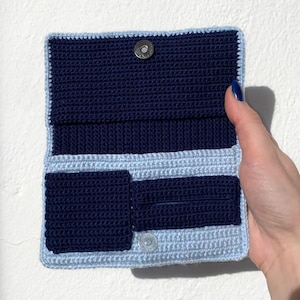 May include: A handmade crochet wallet in shades of navy blue and light blue. The wallet features a rectangular shape with a flap closure and a snap button. The interior has multiple compartments for cards and cash. The crochet stitches are visible, giving it a textured appearance.
