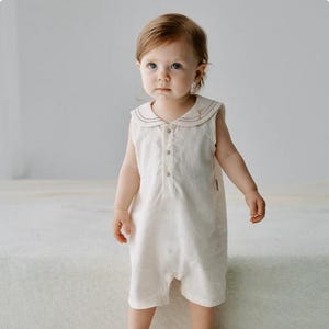 Op de afbeelding: Een cr&egrave;mekleurige babyromper met een matrozenkraag. De mouwloze romper heeft een knoopsluiting aan de voorkant en korte pijpen. De kraag is afgezet met een dunne, donkerdere draad. De baby staat op een lichtgekleurd oppervlak.