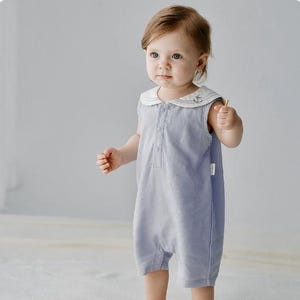 Op de afbeelding: Een lichtblauwe babyromper met een witte matrozenkraag met een geborduurd ankerontwerp. De mouwloze romper heeft een knoopsluiting aan de voorkant en korte pijpen. De baby houdt een klein, dun, geel voorwerp vast.