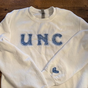 May include: White sweatshirt with the letters "UNC" in blue denim-like fabric across the chest. A small blue heart is on the left sleeve. The sweatshirt is a crew neck style.