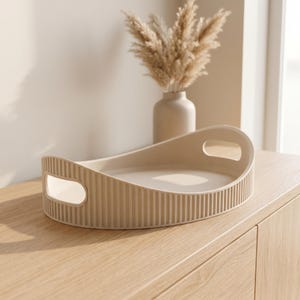Op de afbeelding: Een beige ovaal dienblad met een geribbelde buitenkant en twee geïntegreerde handgrepen aan de zijkanten. Het dienblad heeft een gladde, platte bodem en gebogen wanden, vervaardigd uit een stevig materiaal met een matte afwerking in zandkleur.