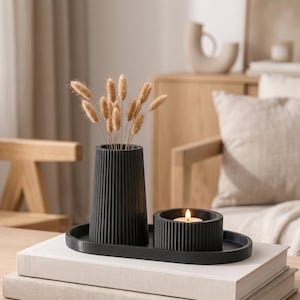 Könnte beinhalten: Eine schwarze, gerippte Vase und ein passender schwarzer, gerippter Teelichthalter mit brennender Kerze, angeordnet auf einem schwarzen ovalen Tablett. Die Vase enthält mehrere hellbraune, getrocknete Gräser, die ein strukturiertes Arrangement bilden.