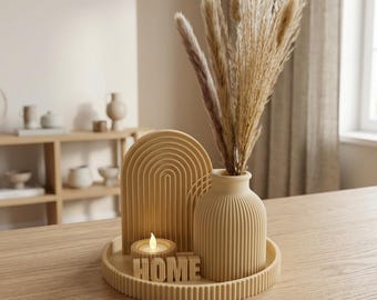 May include: A beige decorative set on a circular ribbed tray, featuring a ribbed vase filled with dried pampas grass, an arched sculpture, a small candle holder with a lit candle, and block letters spelling out the word HOME in beige.