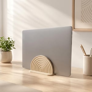 Könnte beinhalten: Ein silberner Laptop, der von einem hellbeigen, halbkreisförmigen Holzständer aufrecht gehalten wird, welcher mit einer Reihe konzentrischer, erhabener Bogenmuster versehen ist. Der Ständer bietet eine stabile Basis in einer neutralen Farbe.