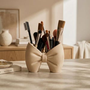 Può includere: Un organizer da scrivania beige a forma di fiocco, realizzato in materiale opaco, contenente vari pennelli per il trucco e un pettine per ciglia. L'organizer presenta un nodo centrale e due anelli simmetrici come base per gli strumenti.