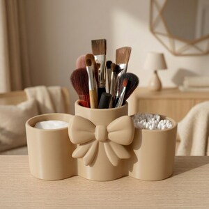 Può includere: Un organizer da trucco beige a tre scomparti con un dettaglio a fiocco centrale. La sezione centrale contiene vari pennelli da trucco con setole marroni e nere, mentre gli scomparti laterali contengono dischetti di cotone e cotton fioc bianchi.