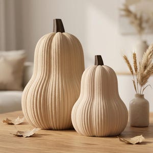 May include: Two decorative pumpkins in a light beige, textured finish with prominent vertical ribs and dark brown stems. The pumpkins are shaped like gourds, with one being taller and larger than the other, both featuring a matte, uniform surface.